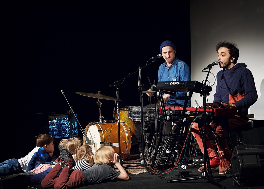 Warsztaty muzyczne dla dzieci — dwóch muzyków gra na keyboards i perkusji, przed sceną na podłodze siedzą i leżą dzieci słuchające koncertu. Zajęcia w ramach programu Katowice Miasto Ogrodów.