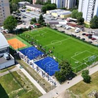 Kompleks boisk przy SP 54 w Giszowcu widziany z lotu ptaka — boisko piłkarskie z zieloną murawą, niebieskie boiska do streetballu i pomarańczowe boisko do siatkówki