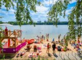 Plaża miejska nad jeziorem w Katowicach — wieża ratownicza MOSiR Katowice, plażowicze opalający się na piasku i kąpiący się w jeziorze, w tle zieleń lasów i osiedlowe wieżowce.