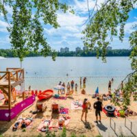 Plaża miejska nad jeziorem w Katowicach — wieża ratownicza MOSiR Katowice, plażowicze opalający się na piasku i kąpiący się w jeziorze, w tle zieleń lasów i osiedlowe wieżowce.