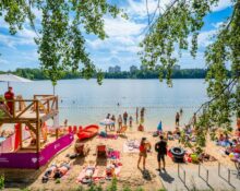 Plaża miejska nad jeziorem w Katowicach — wieża ratownicza MOSiR Katowice, plażowicze opalający się na piasku i kąpiący się w jeziorze, w tle zieleń lasów i osiedlowe wieżowce.